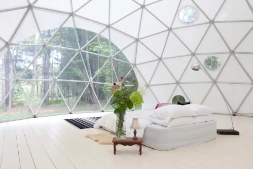 A cozy, modern bedroom inside a geodesic dome, featuring a bed, flowers, and large windows overlooking a forest.
