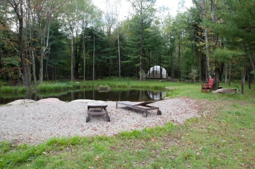 A serene outdoor scene featuring a pond, wooden benches, and a white tent surrounded by trees.