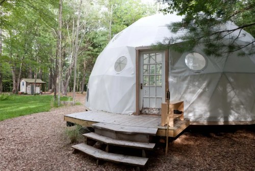 A white geodesic dome with a wooden deck, surrounded by trees and a grassy area, featuring a small shed in the background.