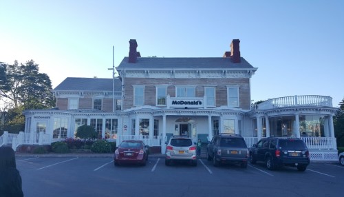 A McDonald's restaurant in a historic building with a porch, surrounded by parked cars and greenery.