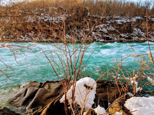 A river with turquoise water flows past rocky banks, surrounded by snow and bare trees in a winter landscape.