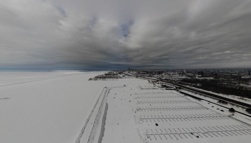 A snowy landscape with a frozen lake, empty parking lot, and a distant city skyline under a cloudy sky.