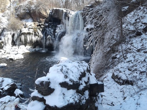 A frozen waterfall surrounded by snow-covered rocks and trees, creating a serene winter landscape.