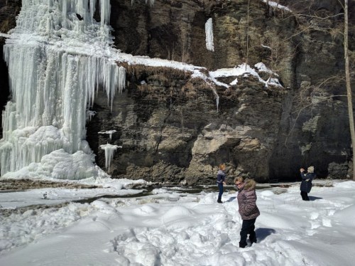 Three people play in the snow near a frozen waterfall and rocky cliff on a sunny winter day.