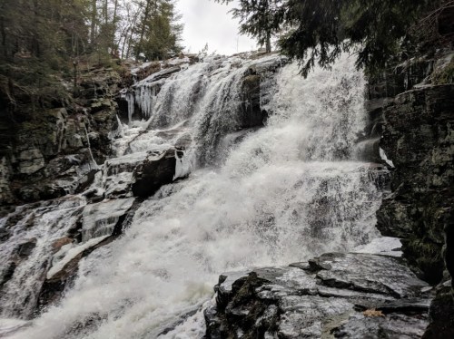 A cascading waterfall flows over rocky terrain, surrounded by trees and mist, creating a serene natural scene.