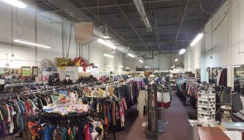 A spacious thrift store aisle filled with racks of clothing and various items for sale. Brightly lit interior.