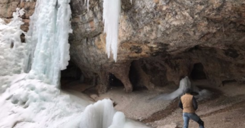 A person stands near icy rock formations and frozen waterfalls in a cave-like setting.