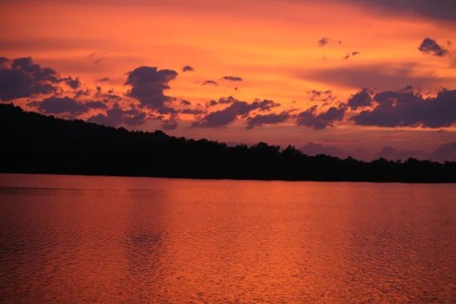 A vibrant sunset over a calm lake, with orange and purple hues reflecting on the water and silhouetted trees in the background.