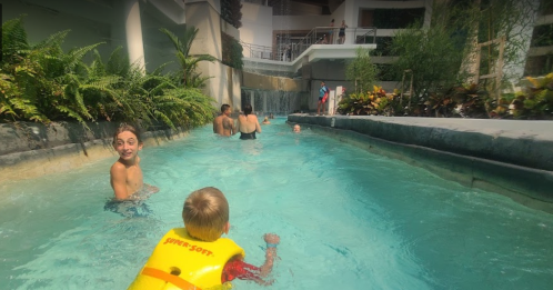 A child in a yellow life jacket swims in a lazy river, surrounded by people enjoying the water. Lush greenery is visible.