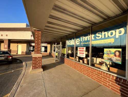 Exterior view of a Value Thrift Shop with large windows displaying signs, located in a shopping plaza.