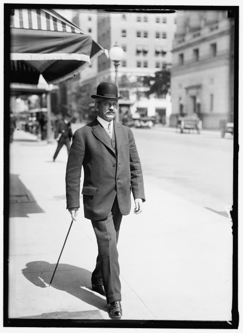 A man in a suit and bowler hat walks confidently down a city street, holding a cane.