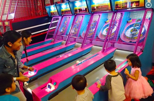 Two adults and three children play a colorful arcade game with pink lanes and balls in a lively setting.