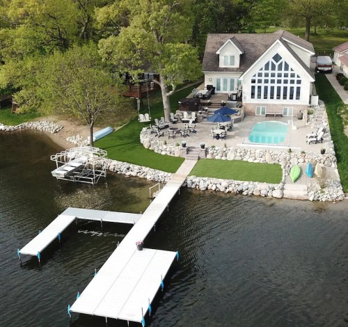 Aerial view of a lakeside home with a pool, dock, and patio surrounded by greenery and water.