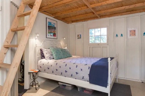 Cozy bedroom with a white bed, blue accents, wooden ceiling, and a ladder leading to a loft area.