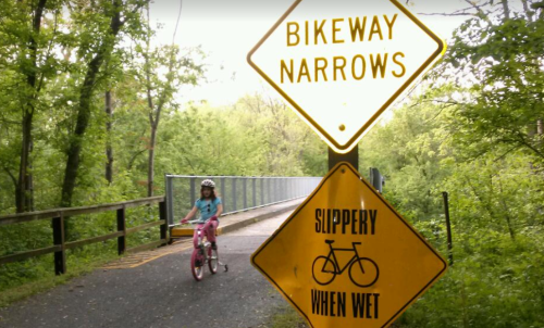 A child on a pink bicycle rides near warning signs for bikeway narrows and slippery conditions when wet.