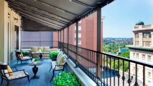 A spacious balcony with seating and plants, overlooking a cityscape under a clear blue sky.