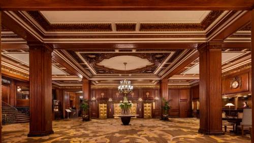 Luxurious hotel lobby with ornate woodwork, a chandelier, and elegant decor, featuring tall columns and plush carpeting.