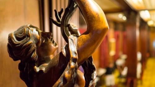 A close-up of a bronze sculpture of a woman, elegantly posed, in a richly decorated interior setting.