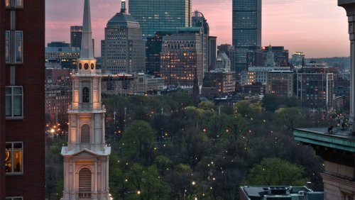 A city skyline at sunset, featuring a historic church steeple and green park areas amidst modern buildings.