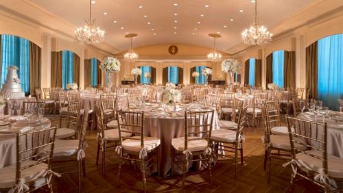 Elegant banquet hall with round tables, gold chairs, floral centerpieces, and chandeliers, illuminated by soft lighting.