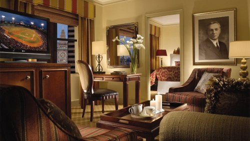 A cozy hotel room with a TV showing a baseball game, a desk, and elegant decor, featuring a portrait on the wall.
