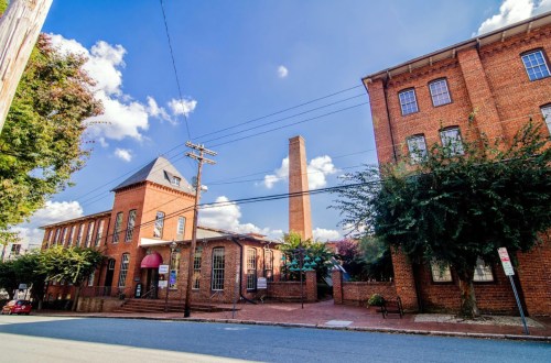 Historic brick buildings with a tall chimney, under a blue sky with fluffy clouds, along a quiet street.