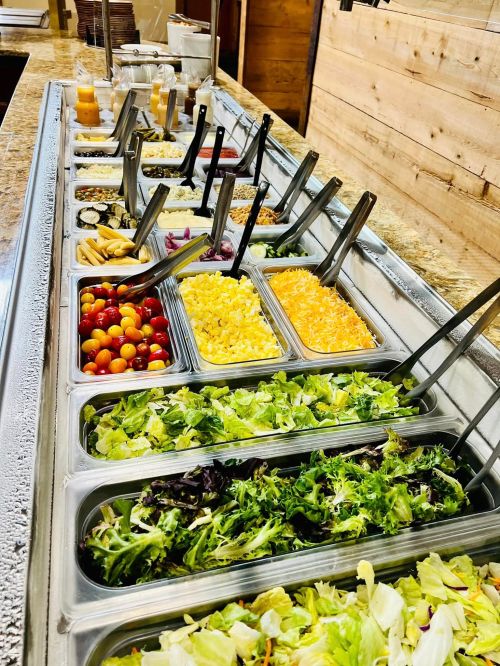 A salad bar featuring various toppings, including tomatoes, cheese, and mixed greens, in stainless steel containers.
