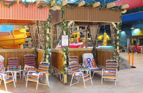 Tropical-themed cabana with colorful lounge chairs and water slides in a vibrant indoor water park setting.