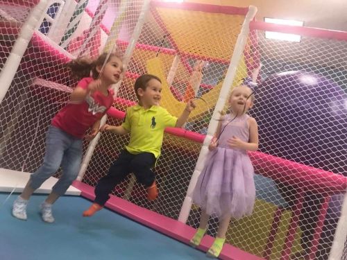 Three children play joyfully in a colorful indoor playground, jumping and smiling amidst netted structures.