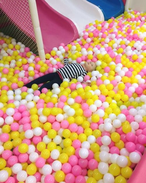 A child in a striped shirt lies among colorful plastic balls in a play area, surrounded by pink and yellow balls.