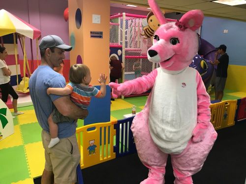 A man holds a toddler as a pink kangaroo mascot waves at them in a colorful play area.