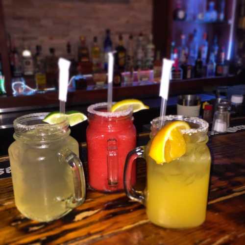 Three colorful cocktails in mason jars with fruit garnishes, set against a blurred bar background.