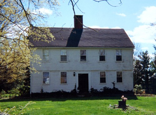 A two-story white house with a black roof, surrounded by greenery and trees under a clear blue sky.