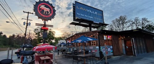 A rustic restaurant exterior with outdoor seating, a neon sign, and a large billboard at sunset.