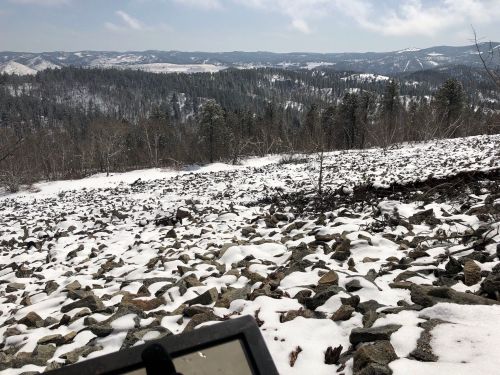 A snowy landscape with scattered rocks, overlooking rolling hills and distant mountains under a cloudy sky.