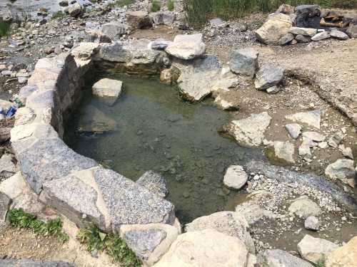 A natural stone pool filled with clear water, surrounded by rocks and gravel, set in a rocky landscape.