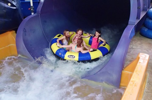 Four children joyfully ride a yellow and blue inflatable tube down a water slide, splashing in the water below.