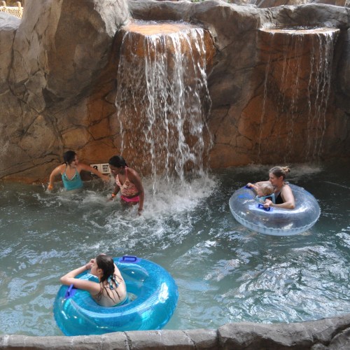 Four people enjoy a pool with a waterfall, two in inner tubes and two playing in the water.