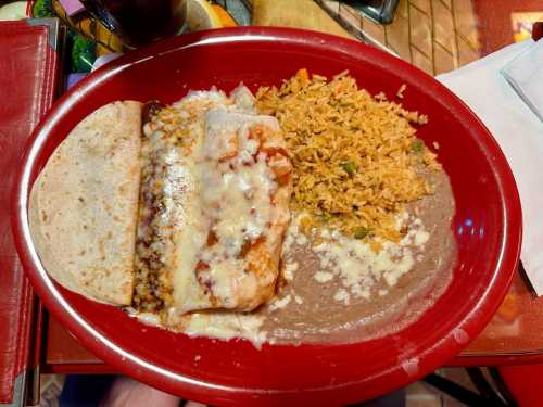 A red plate with a cheese-covered enchilada, rice, refried beans, and a tortilla on a colorful table.