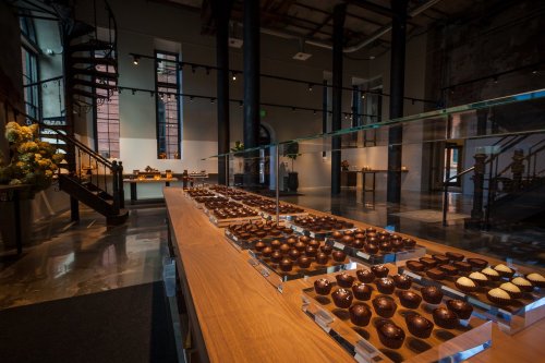 A modern chocolate shop interior featuring a display of assorted chocolates on a wooden table and a spiral staircase.