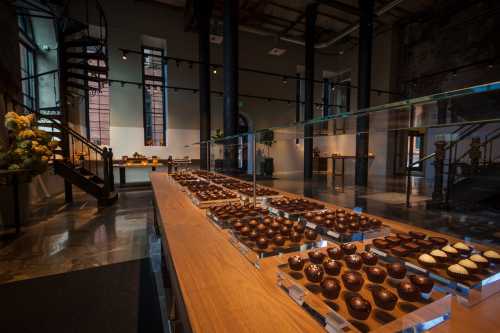 A modern chocolate shop interior featuring a display of assorted chocolates on a wooden table and a spiral staircase.