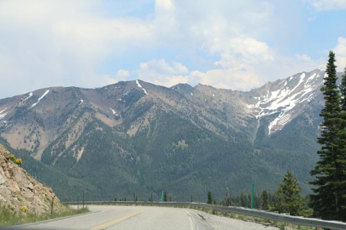 A winding road leads through a mountainous landscape with snow-capped peaks and green forests under a cloudy sky.