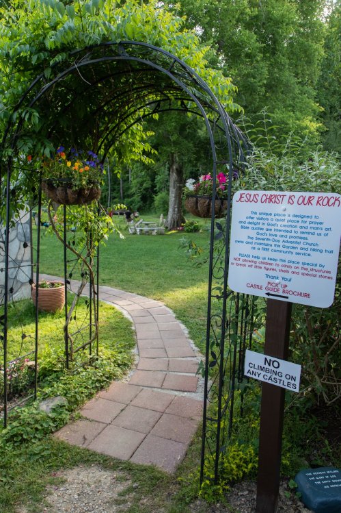 A garden archway adorned with flowers leads to a path, with a sign about a church and a warning against climbing.