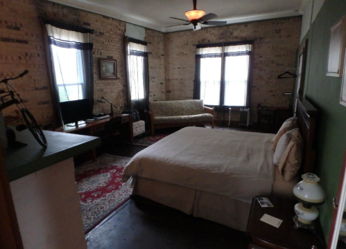 Cozy bedroom with a bed, sofa, TV, and bike, featuring brick walls and large windows with sheer curtains.