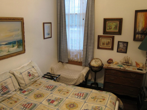 Cozy bedroom with a patterned bedspread, paintings on the walls, a globe, and a small seating area by the window.