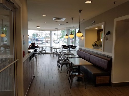 A modern café interior with wooden floors, tables, and chairs, featuring large windows and a cozy seating area.