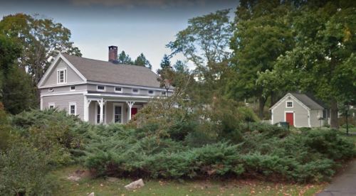 A gray house with a porch and red doors, surrounded by greenery, with a smaller building nearby.