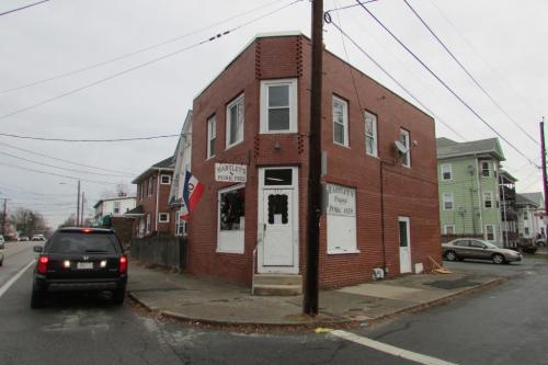 A brick corner building with a sign for a pizza shop, surrounded by power lines and parked cars on a cloudy day.