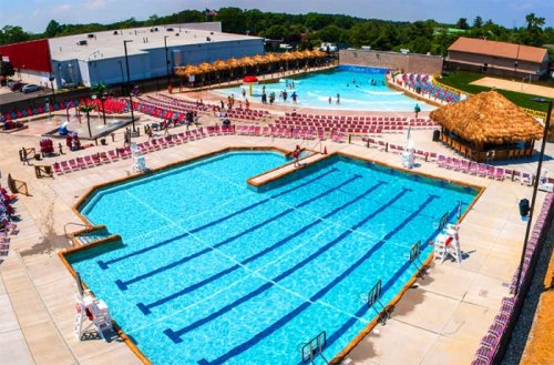 Aerial view of a large outdoor pool area with multiple pools, lounge chairs, and a tiki hut, filled with people enjoying the sun.