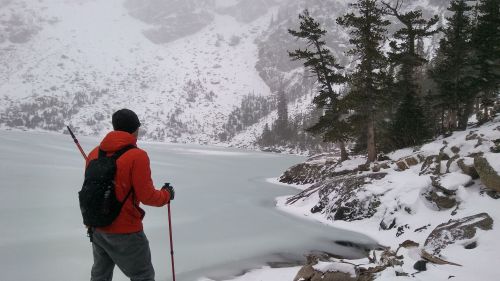 A person in an orange jacket stands by a frozen lake surrounded by snow-covered mountains and trees.